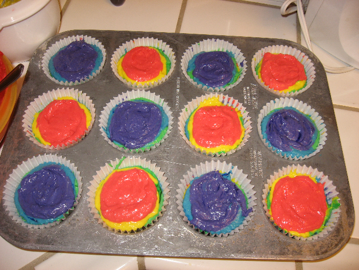 Multiple colors of cupcake batter in twelve cupcake liners in a metal cupcake pan