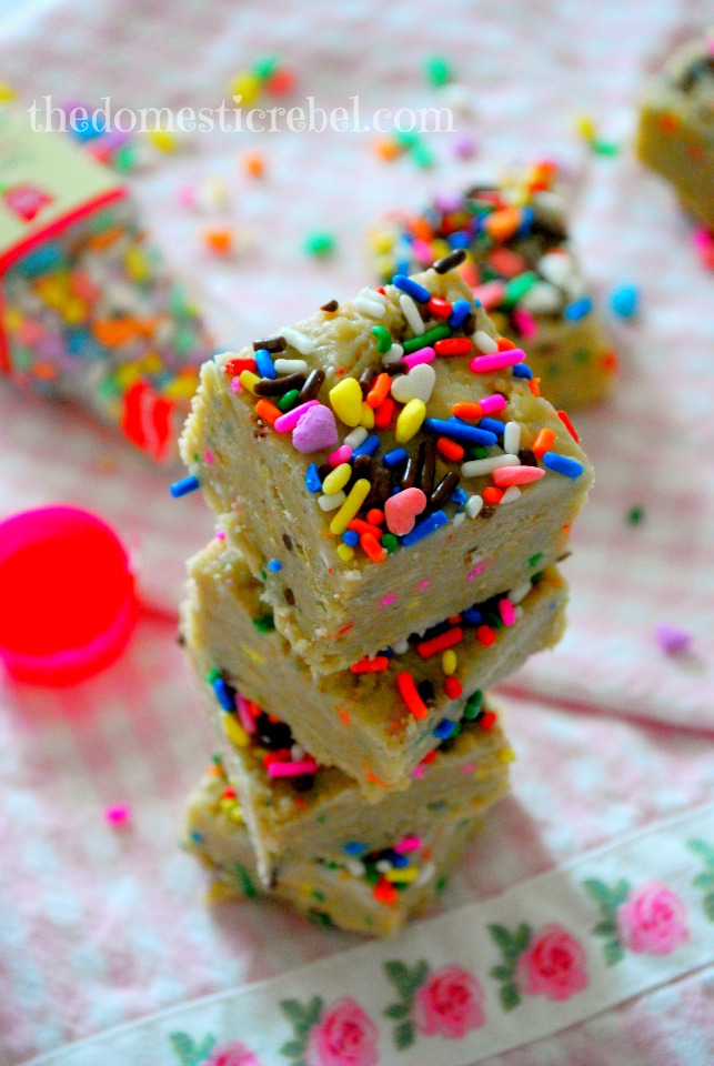 no bake cake batter squares stacked on a pink background with sprinkles in the background