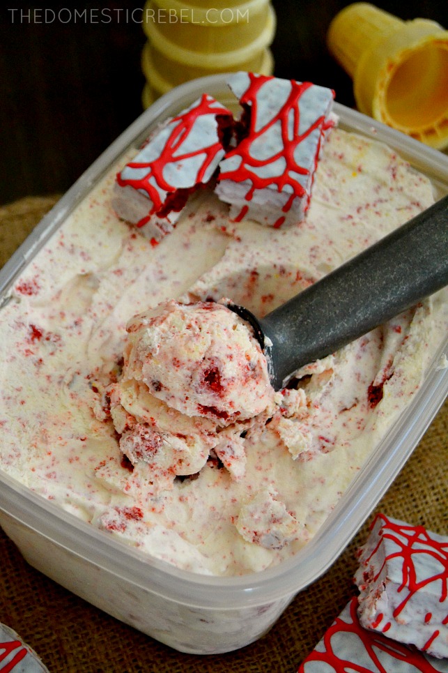 red velvet cheesecake ice cream in a container with a metal ice cream scoop and cones in background