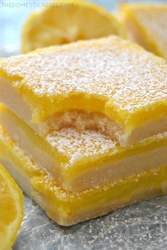 Closeup of Lemon Bars stacked on parchment with bite missing and lemon wedges