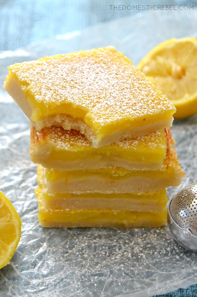Lemon Bars stacked on parchment with lemon wedges and powdered sugar dusting tool