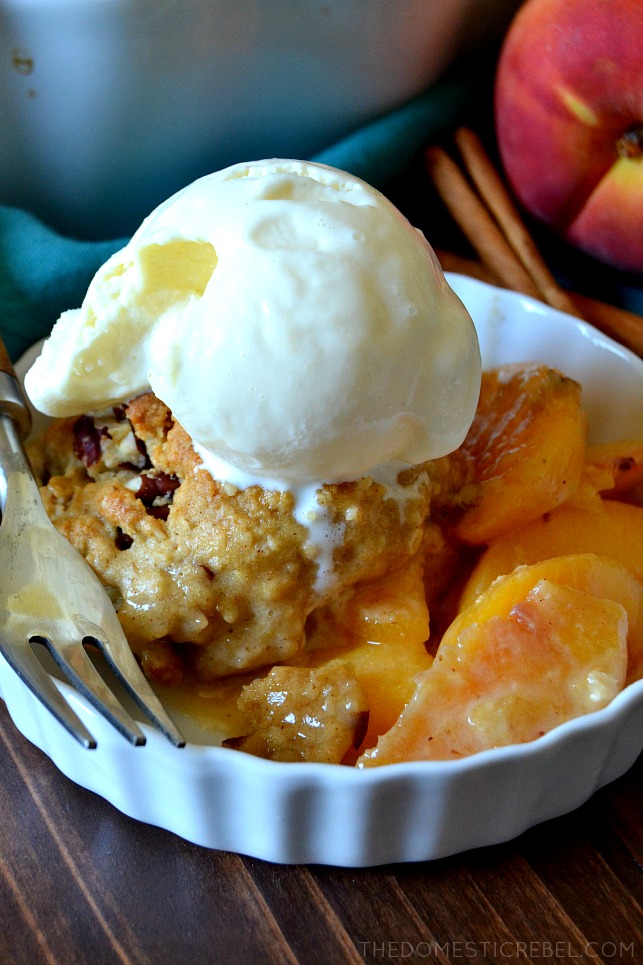 Oatmeal cookie peach cobbler topped with ice cream in a white ramekin next to a fork