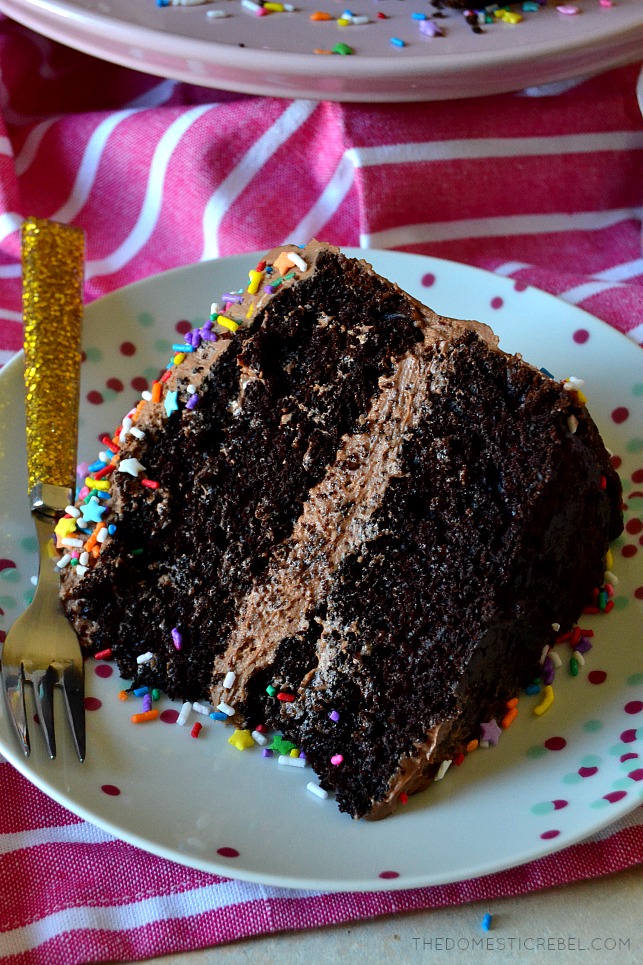 Slice of chocolate layer cake on a white plate next to a gold, sparkly fork