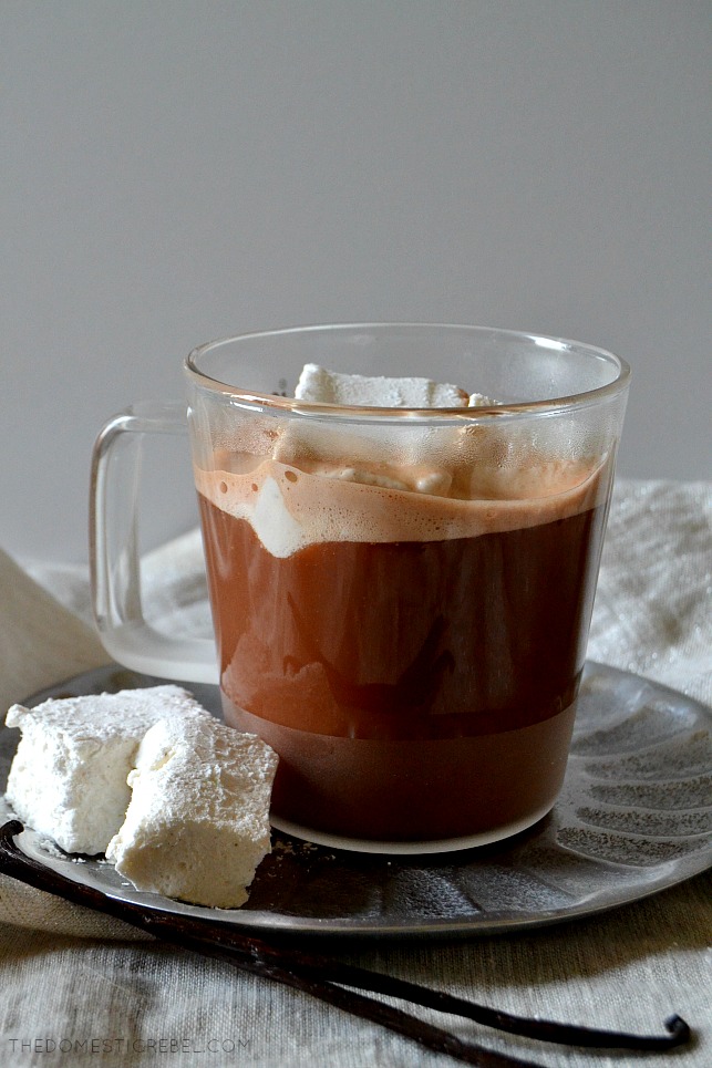 A clear mug of hot chocolate with a marshmallow in it, with marshmallows and vanilla bean around the mug