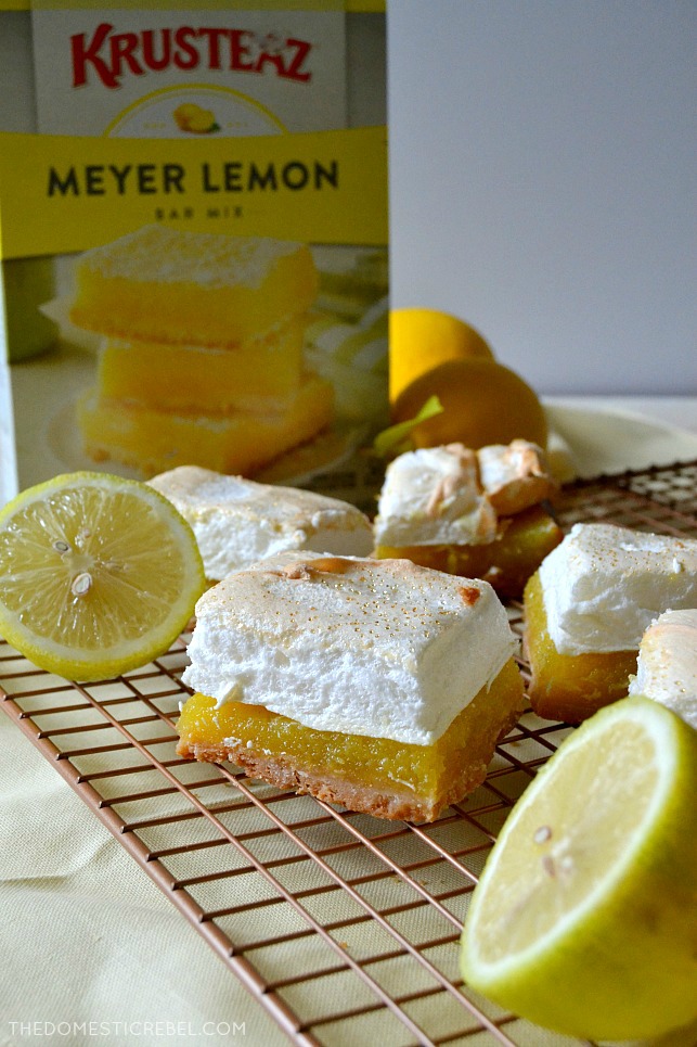 A few lemon meringue pie bars on a wire rack with halved lemons, all in front of a box of Krusteaz Meyer Lemon Bar Mix