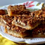 pecan pie bars arranged in a stacked formation on a white plate on top of a floral dish towel
