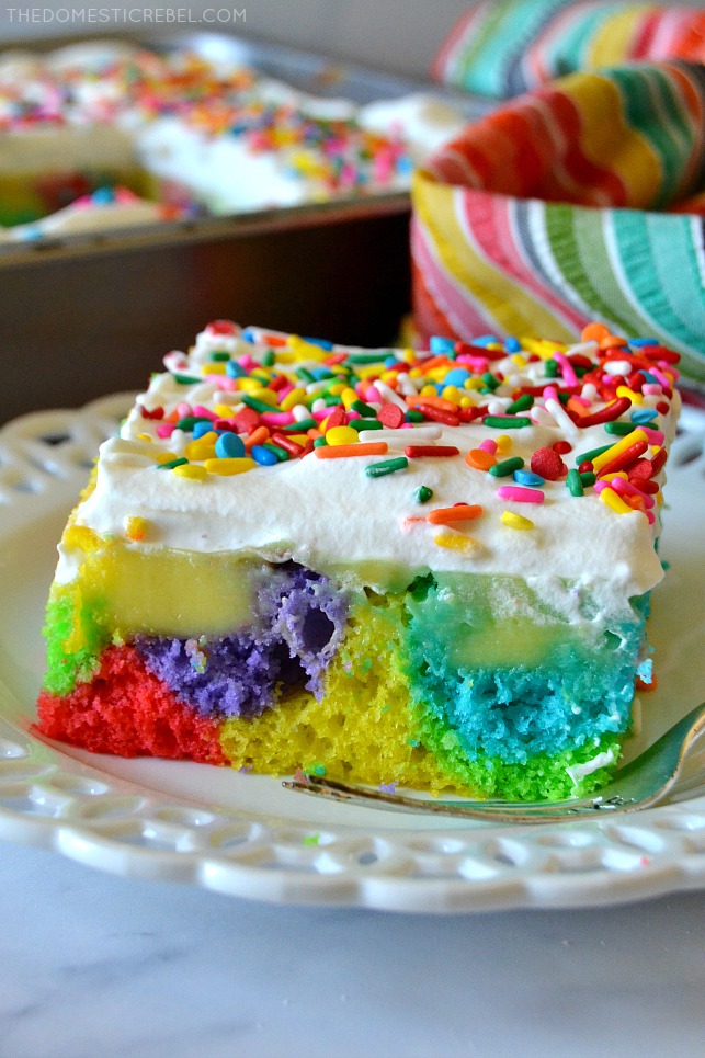 Closeup of Rainbow Poke Cake slice on white plate with rainbow fabric in background