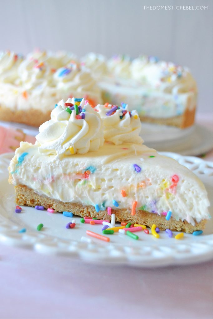 a side profile of a piece of cake batter cheesecake on a white scalloped plate with sprinkles on it.