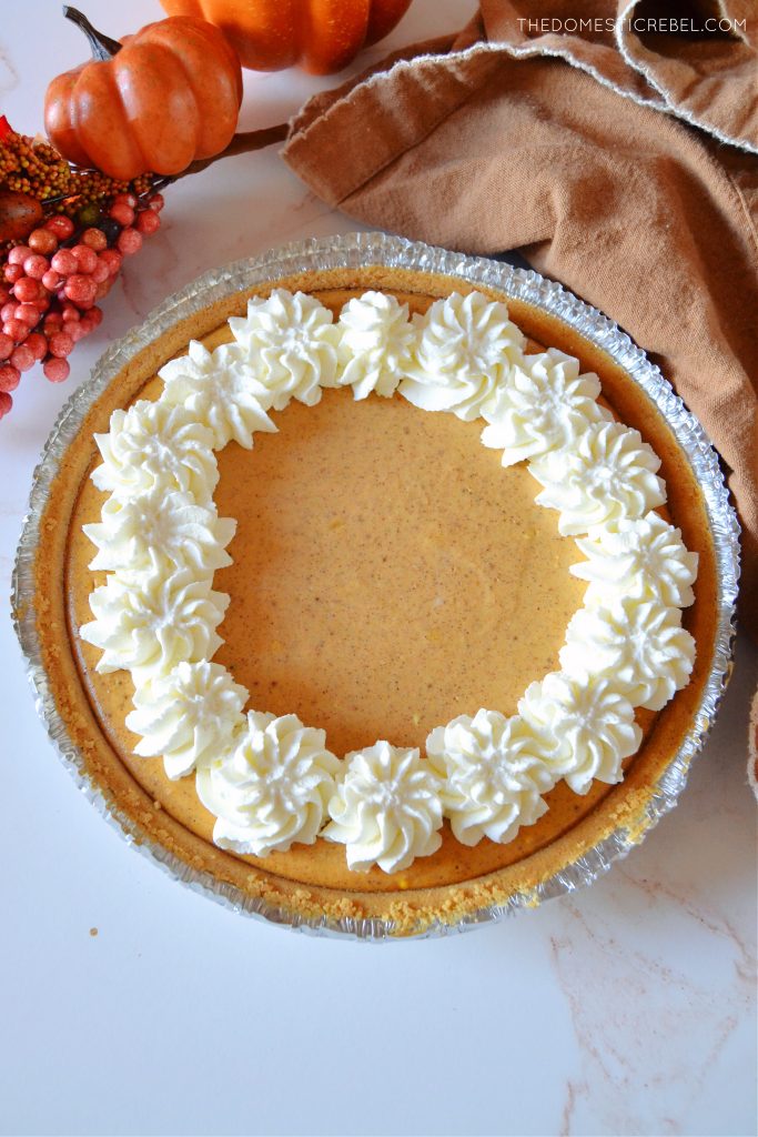 an aerial shot of an entire pumpkin pie cheesecake in the pan with a brown towel and some faux pumpkins and nature elements surrounding it on a white marble background