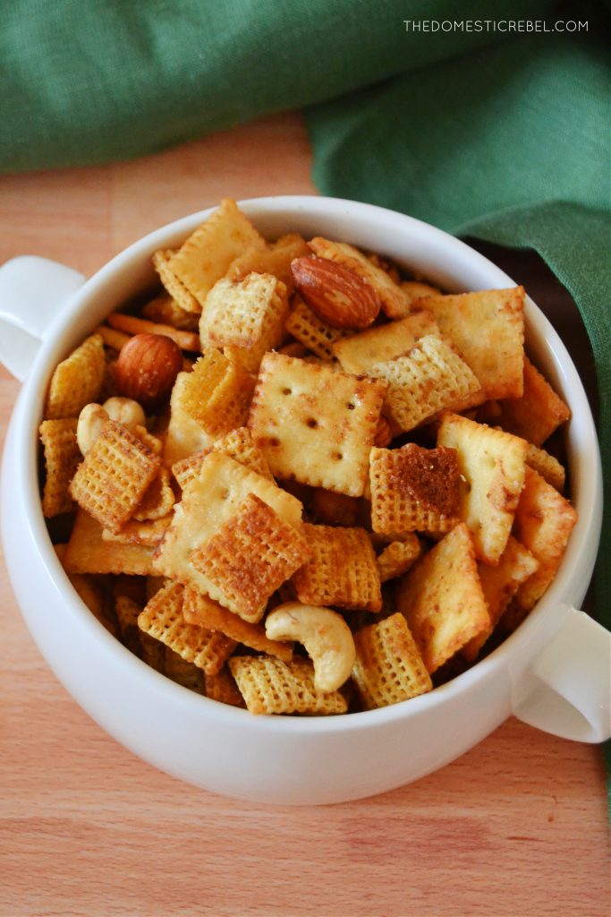an aerial shot of chex mix in a handled white bowl on a wooden board with a green towel in the background