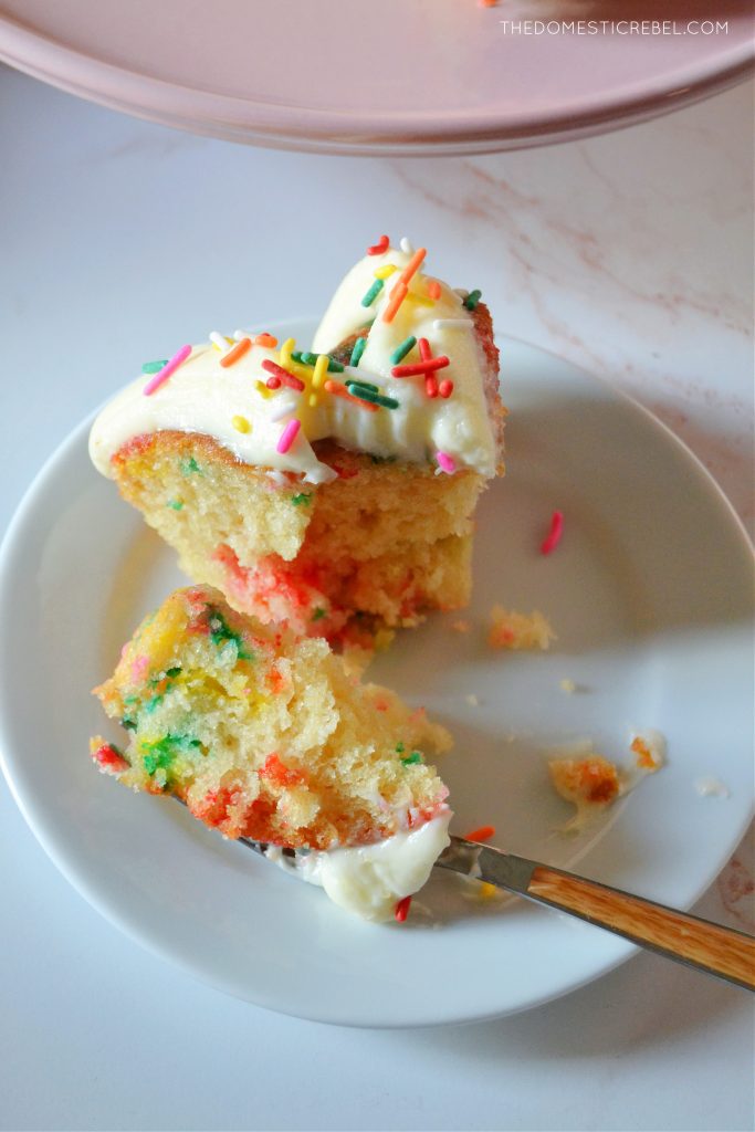 an aerial shot of a piece of funfetti bundt cake with a bite missing out of it. A silver and wooden fork rests on the plate with a chunk of cake on the fork.