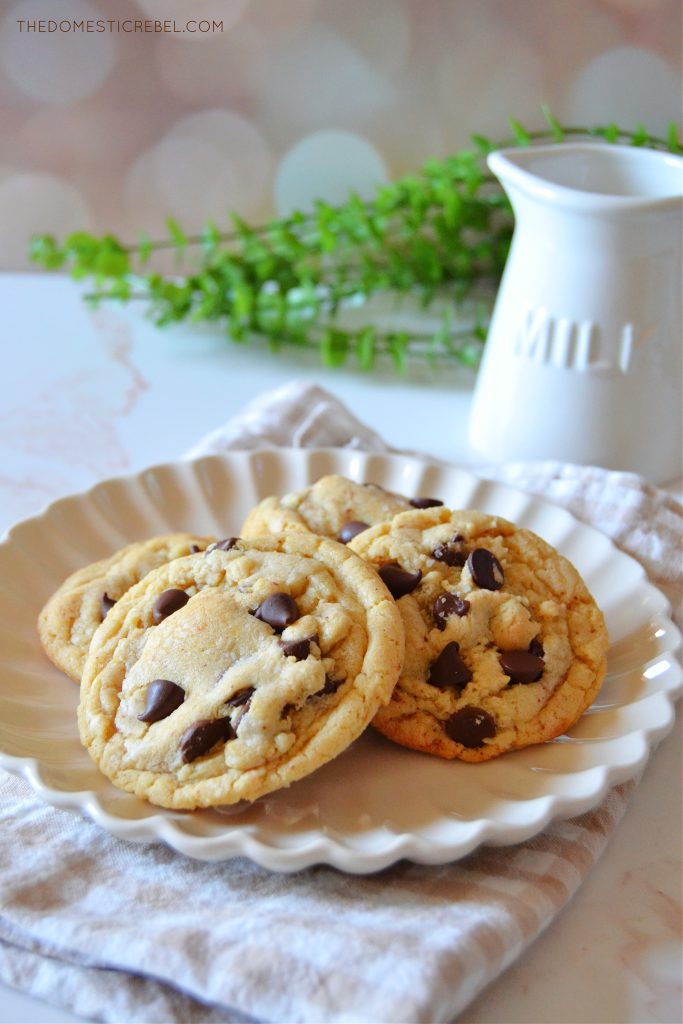 a white scalloped plate full of brown butter chocolate chip cookies sitting on top of a beige gingham napkin on a marble board. A milk jug and a piece of greenery are in the background.