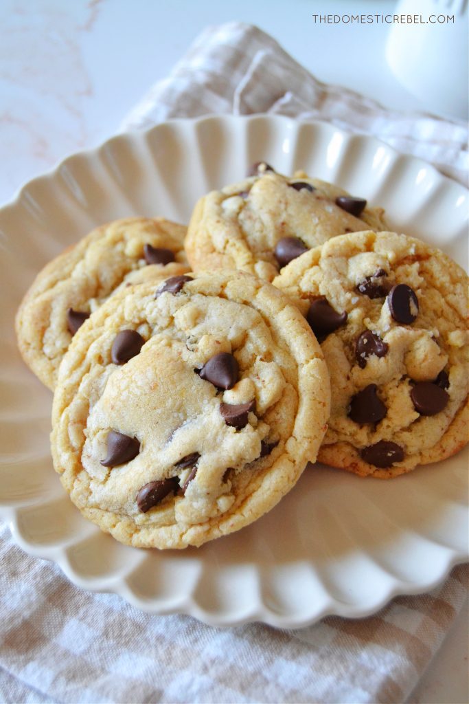 a white scalloped plate filled with four brown butter chocolate chip cookies sitting on a beige gingham napkin