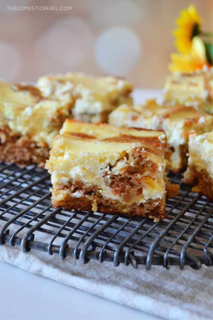 an arrangement of carrot cake cheesecake bars on a black wire rack sits against a light spotted background. some yellow flowers are off to the right in the background.