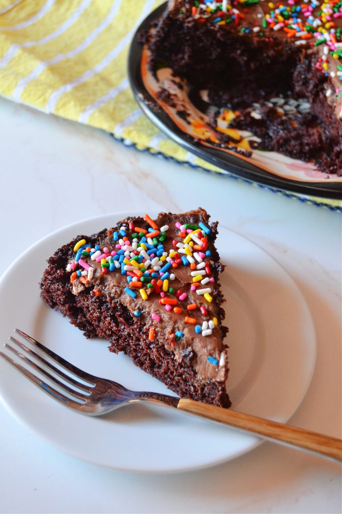 an aerial shot of a slice of chocolate snack cake on a small white plate with a wooden fork resting on the plate. a larger plate of the cake sits in the background on a yellow striped dish cloth.