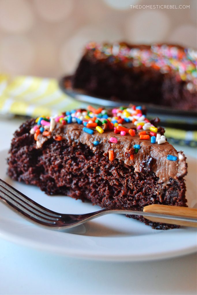 a closeup shot of a slice of chocolate snack cake on a white plate with a fork sitting on it. more cake is sitting in the background on a yellow cloth.