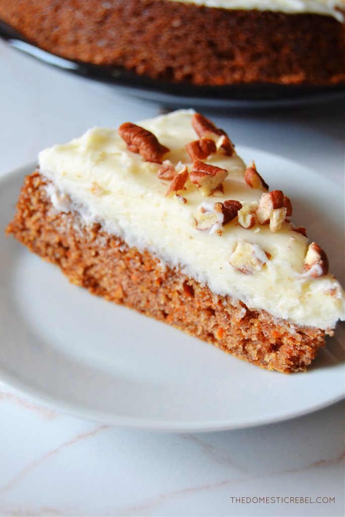 a slice of carrot snacking cake sits on a petite white plate. the larger carrot cake rests in the background on a black plate.