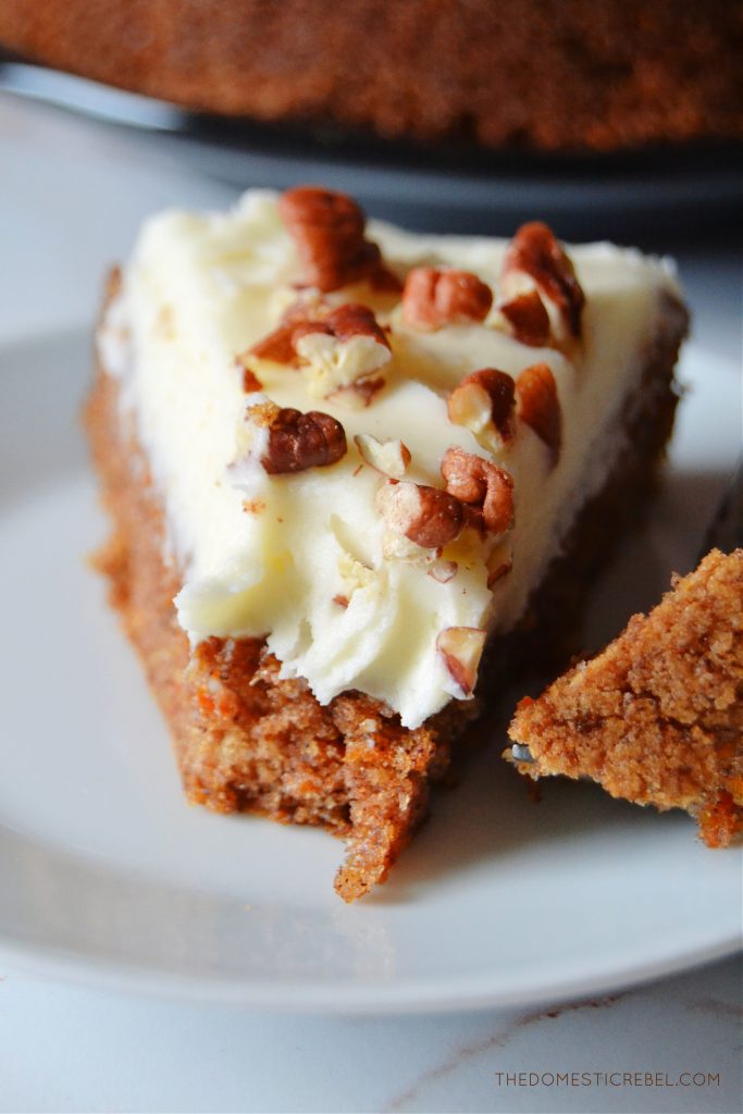 a closeup slice of carrot cake with a bite missing from the slice. it is sitting on a white plate.