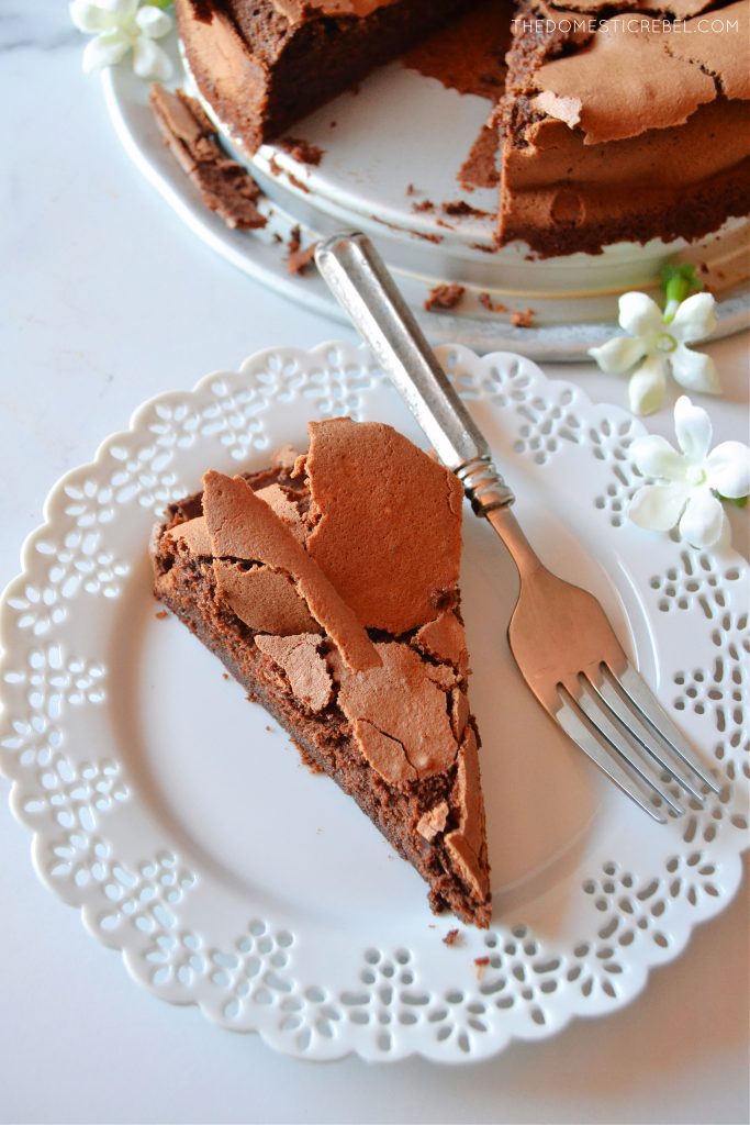 an aerial shot of a piece of flourless chocolate cloud cake on a white scalloped plate. a fork rests on the plate and two little white flowers are sitting near the plate.