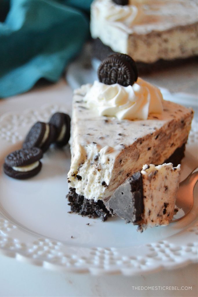 a slice of oreo cheesecake sits on a white lacy plate. a fork is on the plate with a bite of the cheesecake on the fork. 