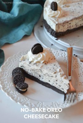 a single slice of oreo cheesecake rests on a white lacy plate with a silver and wood fork and three tiny oreo cookies.