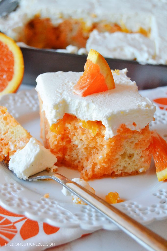 a slice of orange creamsicle poke cake on a white scalloped plate with a fork. there is a bite missing from the cake.