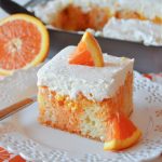 a single slice of orange creamsicle poke cake sits on a scalloped white lace plate with a fork and an orange slice.
