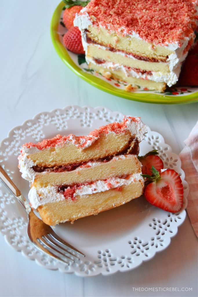 an aerial shot of a slice of strawberry shortcake icebox cake on a white lacy plate with a fork. it sits next to the whole cake on a green plate.