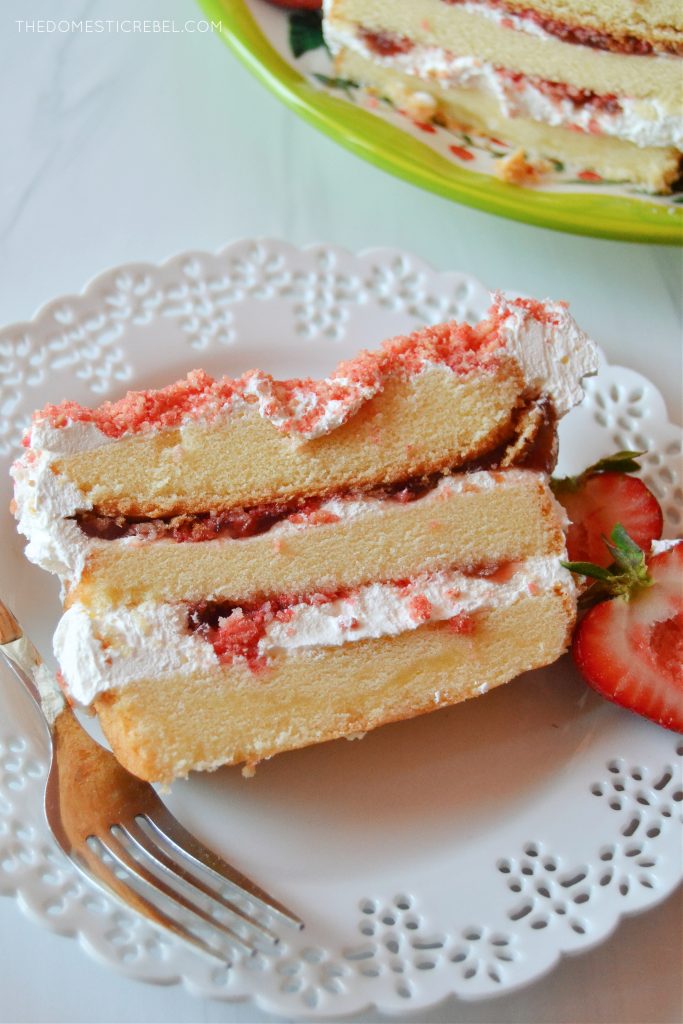 a slice of strawberry shortcake icebox cake sits on a white lacy plate with a fork and a halved strawberry.