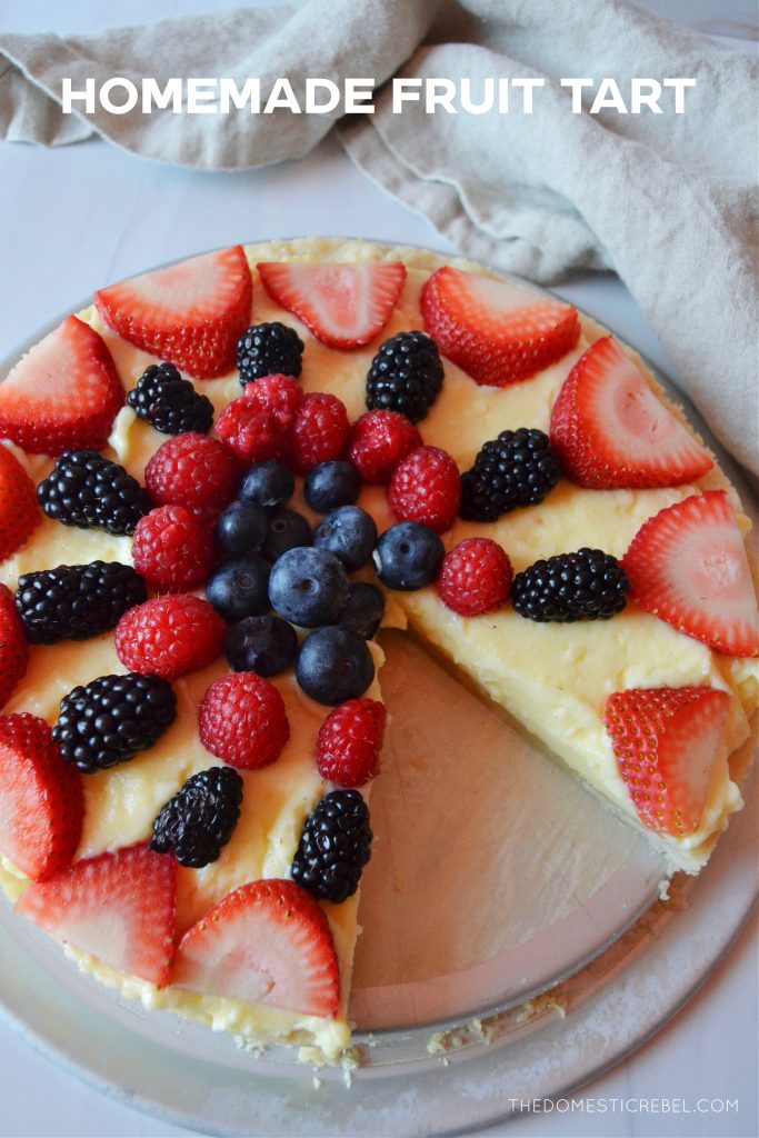 a fruit tart with a slice missing from it sits on a white board with a linen napkin resting beside it.