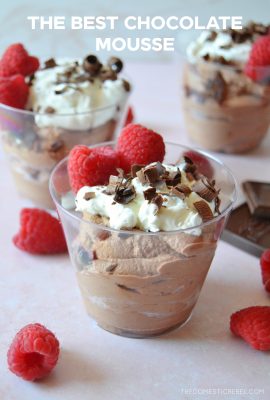 three glasses of chocolate mousse parfaits sit on a light pink background with fresh raspberries scattered around them.