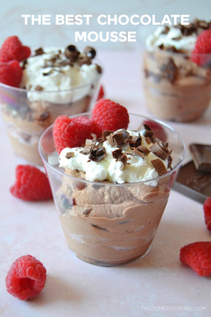 three cups of chocolate mousse sit in the photo on a pink background with fresh raspberries scattered around them. 