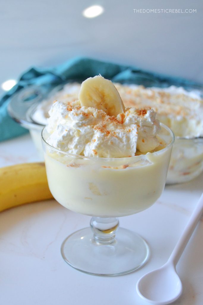 a small trifle dish filled with banana pudding sits on a white marbled board with a plastic white spoon next to it and a banana behind it.