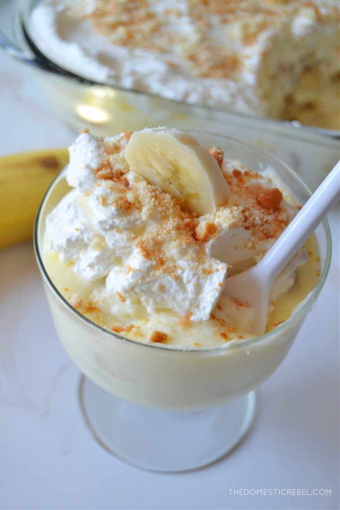 an aerial shot of a trifle dish filled with banana pudding. a white plastic spoon is digging into the banana pudding.