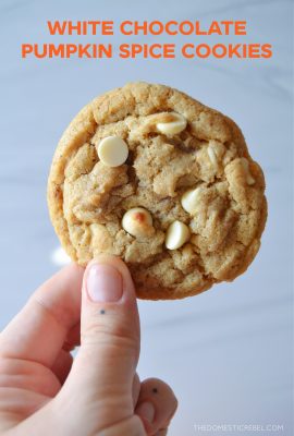 a human hand holds up a white chocolate pumpkin spice cookie against a light colored background.