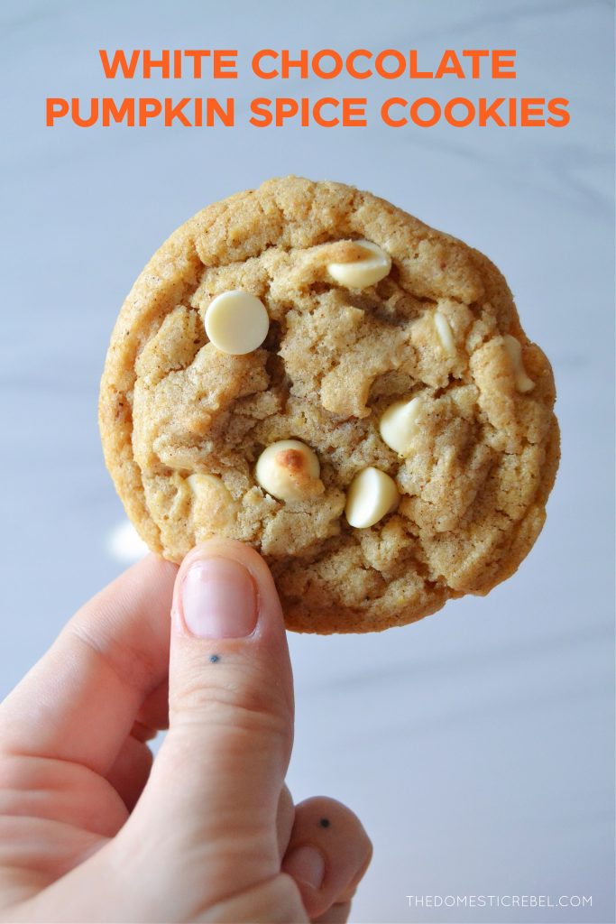 a hand is holding a white chocolate pumpkin spice cookie up to the camera against a light colored marbled background. text on the photo says "white chocolate pumpkin spice cookies."