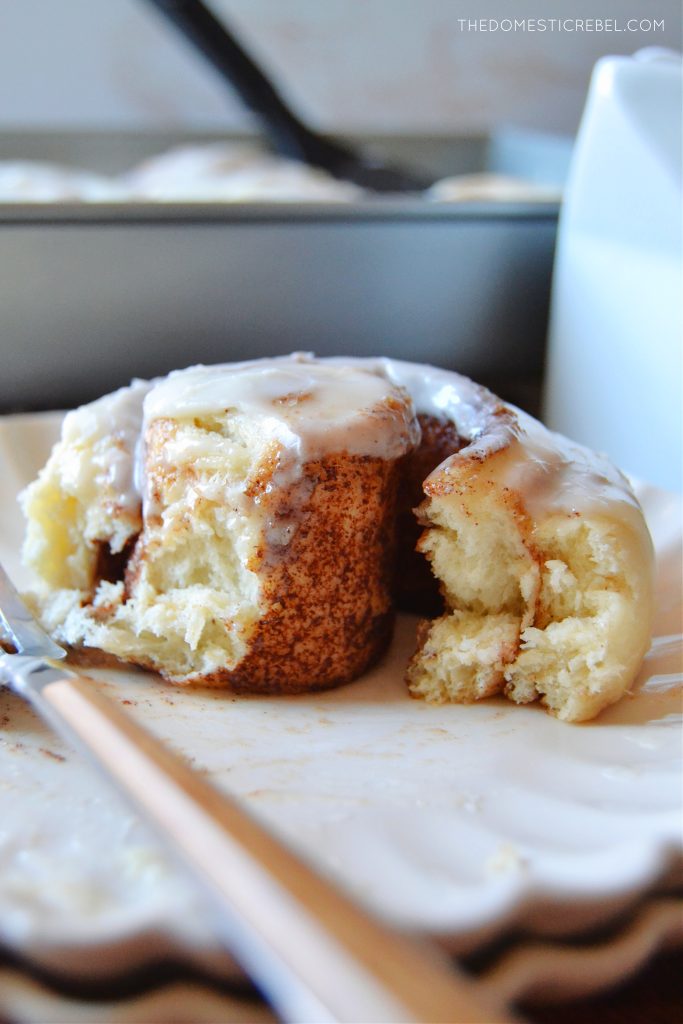 a cinnamon roll slightly unwound from its spiral to show the inside of the roll. it sits on a scalloped white plate with a wooden fork.