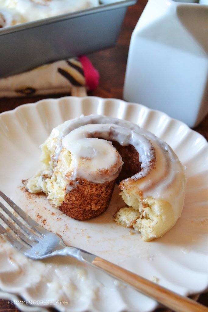 a half-eaten cinnamon roll on a white scalloped plate with a wooden fork. a ceramic milk carton sits in the background.