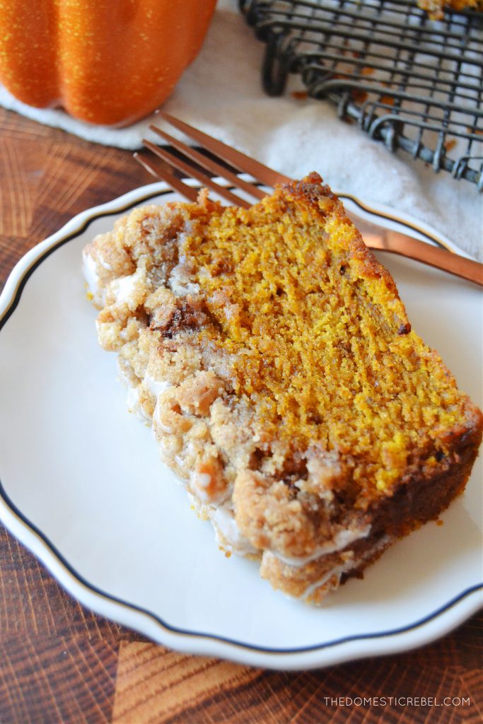 a slice of pumpkin streusel bread on a white scalloped plate with a rose gold fork. the plate is on a wooden cutting board with a pumpkin in the background.