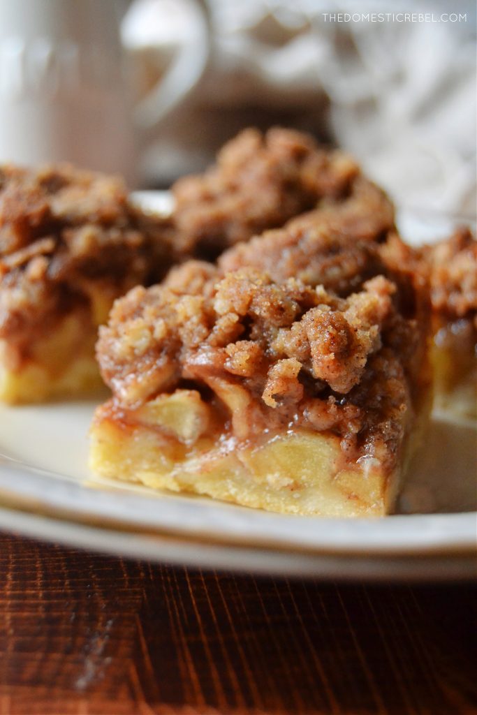 a closeup photo of a dutch apple pie bar on a white plate. a couple other pie bars are in the background.