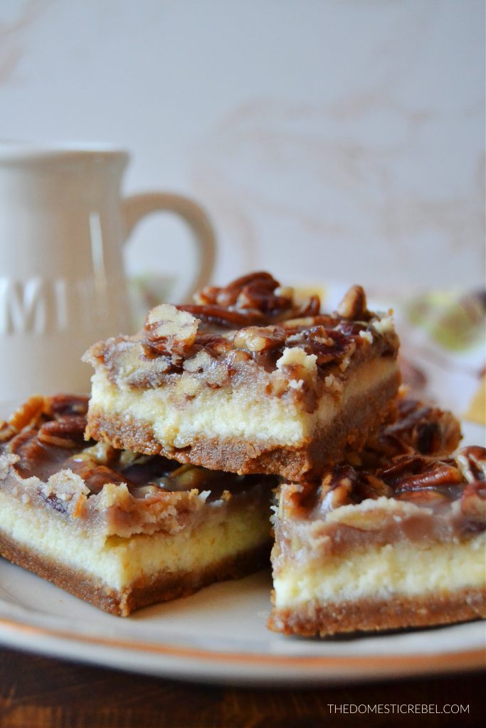 a trio stack of pecan pie cheesecake bars on a gold-rimmed plate. a milk jug is in the background. 