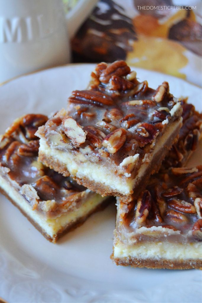 pecan pie cheesecake bars stacked and arranged on a gold-rimmed plate. they're sitting on a floral fall-themed dish towel with a milk jug in the background.