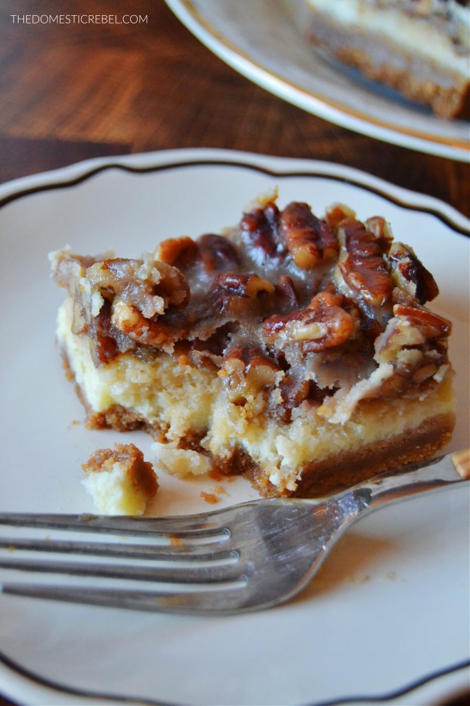 a pecan pie cheesecake bar sits on a black and white plate with a metal fork. a bite has been taken out of the bar.