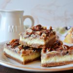 three pecan pie cheesecake bars stacked on a little gold and white plate against a white and wooden background.