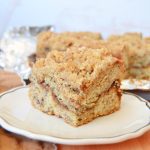 a piece of banana bread coffee cake on a white plate. the plate is on a brown cutting board.