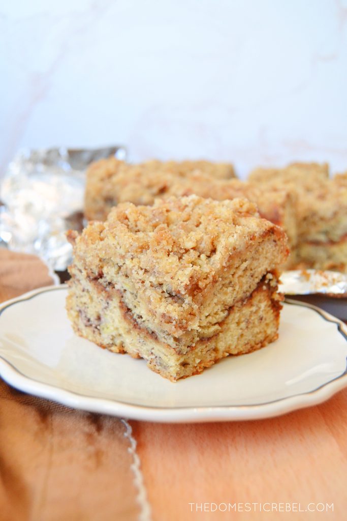 a single piece of banana bread coffee cake sits on a white plate with black trim. the plate is on a wooden surface with a foil-lined pan of more coffee cake in the background.