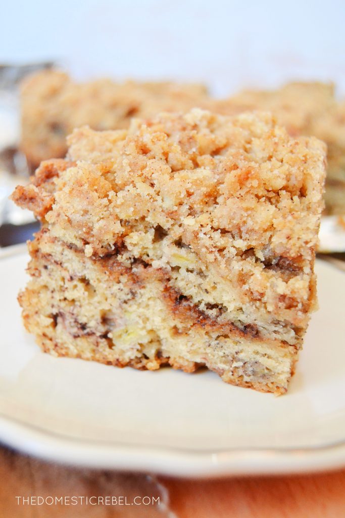 a close-up shot of a piece of banana bread coffee cake on a white scalloped plate. the plate sits on a wooden cutting board with more banana bread coffee cake in the background on a piece of foil.
