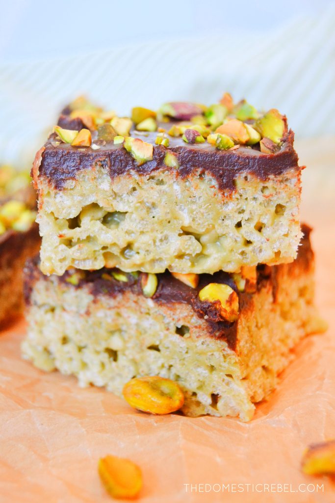 a stack of two dubai-style rice krispy treats sit on brown wrinkled parchment paper. a scattering of shelled pistachios are also on the paper in front of the stack.