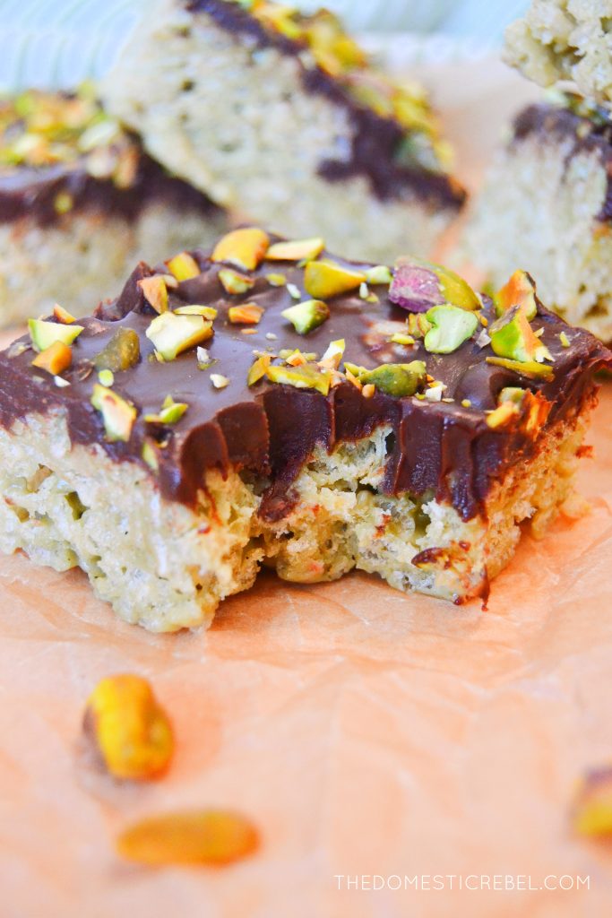 a dubai chocolate rice krispy treat sits on a piece of brown parchment paper, surrounded by other squares of rice krispy treats. the treat in the foreground has a bite taken from it.