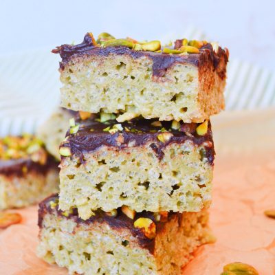a trio stack of dubai rice krispy treats sits on a piece of brown parchment. a small scattering of shelled pistachios are around the stack.